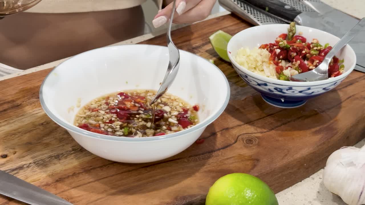Hands prepare a spicy Thai dipping sauce by adding fish sauce, chopped chili, garlic, and lime juice in a well-lit kitchen with steady close-up shots