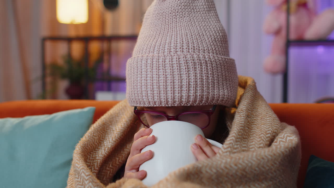 병든 유아 소녀는 모자를 입고 카레드에 싸여 혼자 앉아 소파에서 차가움으로 떨며 뜨거운 차를 마시고 있습니다.