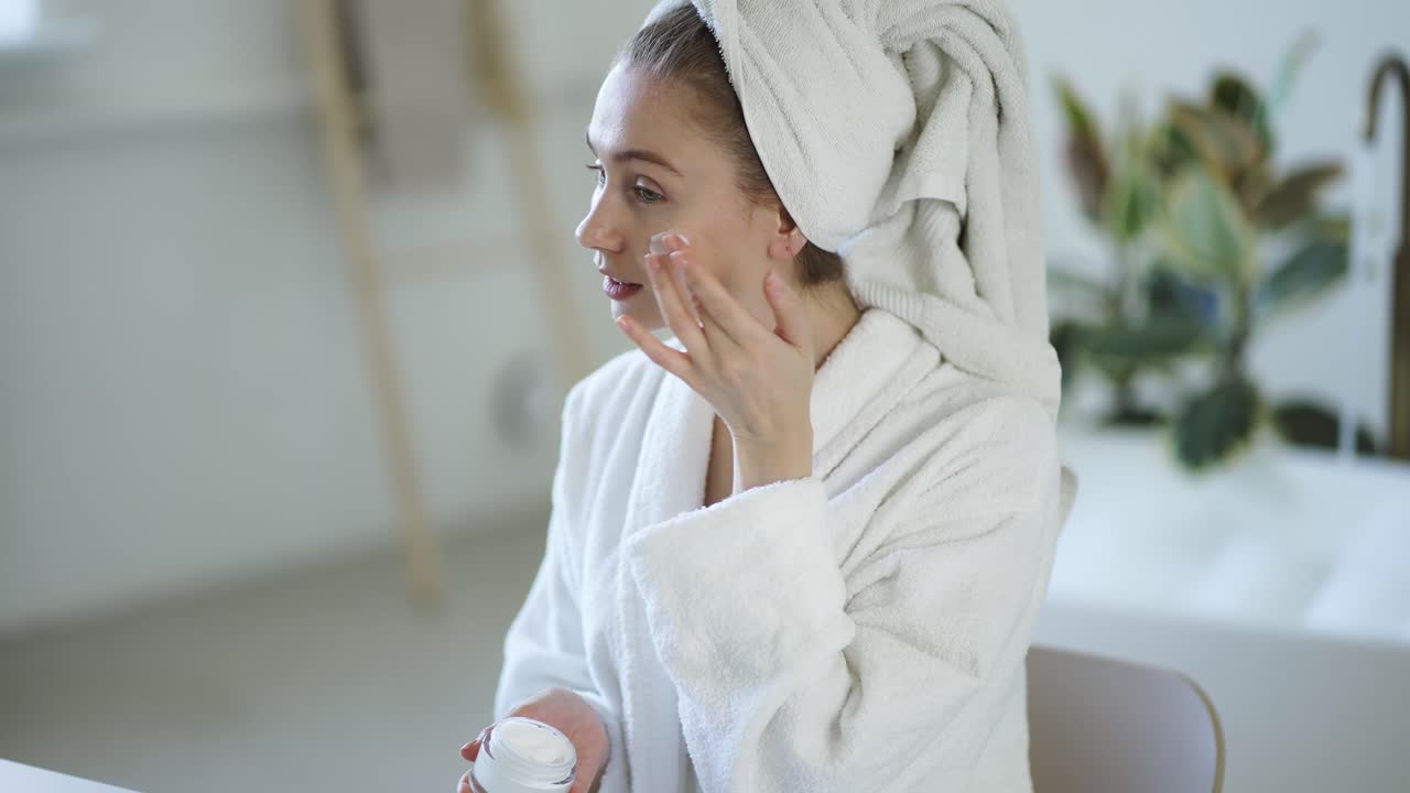 Young woman in white bathrobe and with towel on head applies moisturizer cream on her face