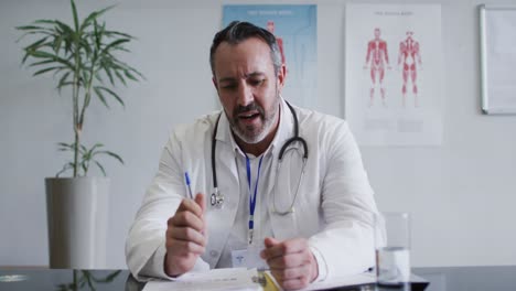 Caucasian-male-doctor-at-desk-talking-and-gesturing-during-video-call-consultation