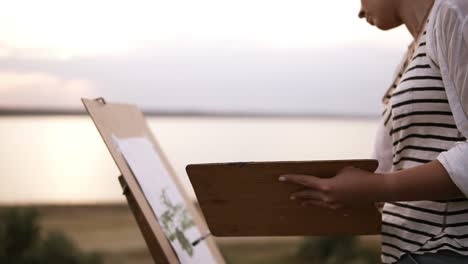 side view of a lovely woman drawing outdoors on the meadow using an easel and palette. blurred lake view on the background