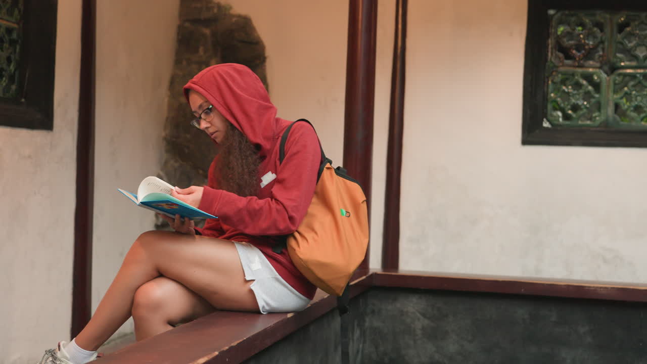 Jovem mulher lendo em uma saliência de pedra, moletom vermelho e mochila laranja, folhagem desfocada em primeiro plano, vista de perfil com diário azul aberto, vibração de viajante relaxado, luz solar da tarde lançando sombras suaves