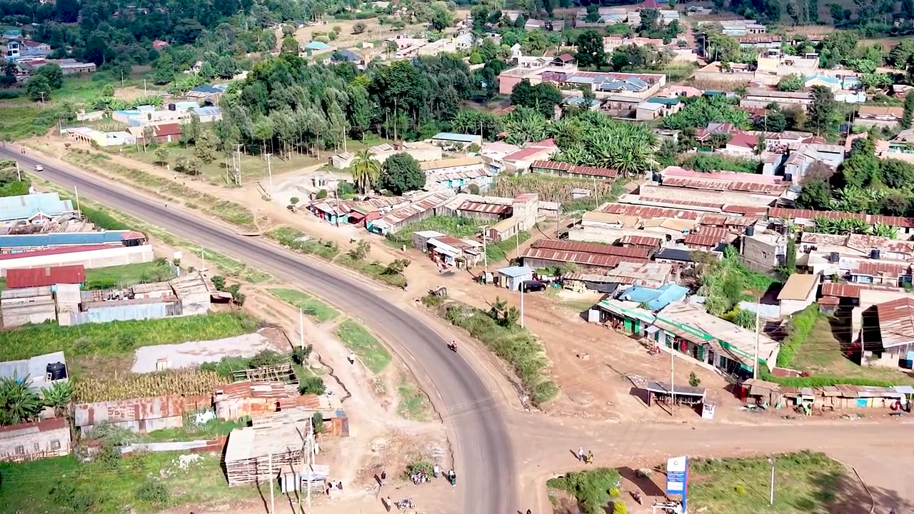Una vista cinematografica del Kenya rurale che mostra insediamenti informali con tetti di lamiera arrugginiti, case logore, sentieri polverosi del villaggio e la vita quotidiana della comunità.