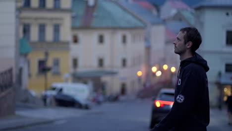 Young-man-with-black-Hoodie-crossing-the-street-in-an-old-historical-town-Banska-Stiavnica-in-Slovakia,-Europe-during-a-blue-hour-at-dusk,-surrounded-by-historical-buildings