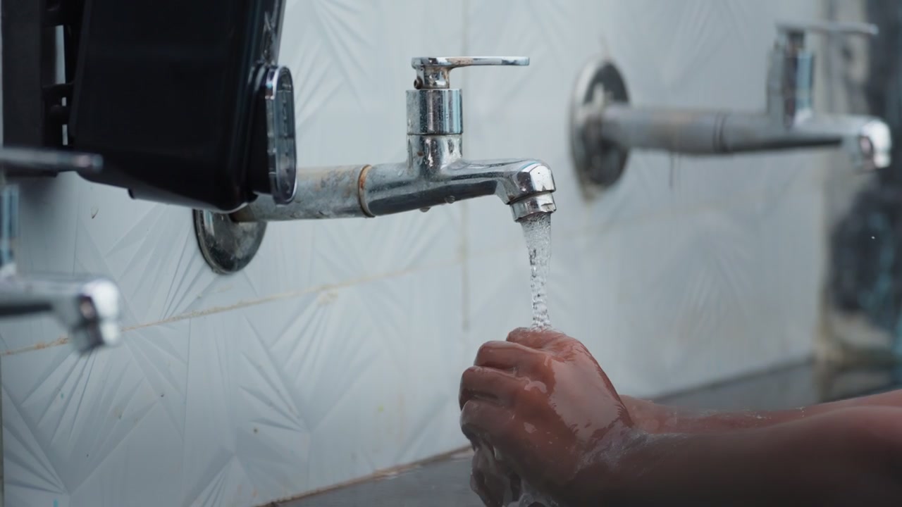 Un bambino si lava le mani sotto un rubinetto cromato in un lavabo semplice in un ambiente scolastico rurale. Il semplice rubinetto a parete rilascia acqua su mani a coppa nella routine di igiene quotidiana di uno studente.