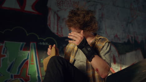 relaxed teenage man resting after riding at skate park with graffiti wall.