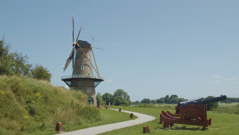 Aufkippen-Traditioneller-Windmühlenflügel,-Die-Sich-Im-Wind-Drehen,-Mit-Alten-Kanonen-Im-Vordergrund