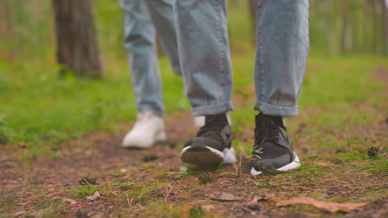 Close-up leg view of two individuals in blue jeans, one in black sneakers and the other in white sneakers, standing on a forest trail, both moves playfully, as if dancing