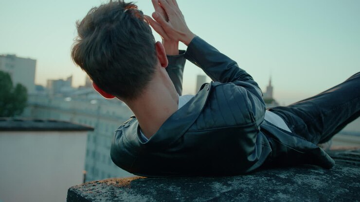 Uomo sdraiato sulla schiena, che indossa una giacca di pelle nera, durante l'ora d'oro al tramonto, con lo skyline della città sullo sfondo, in contemplazione.