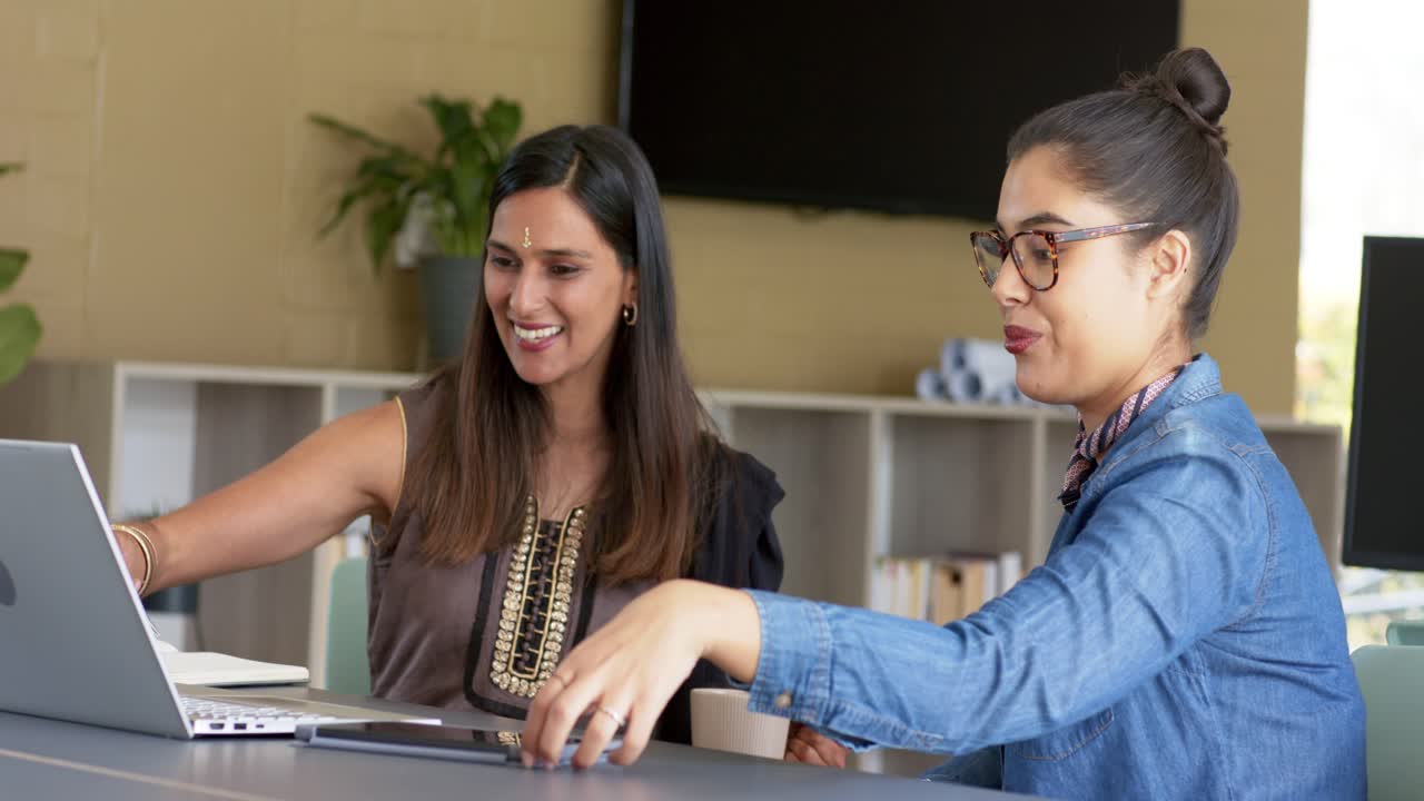 Colegas diversas no escritório apontando para o laptop, conversando, oferecendo tablet e revisando conteúdo