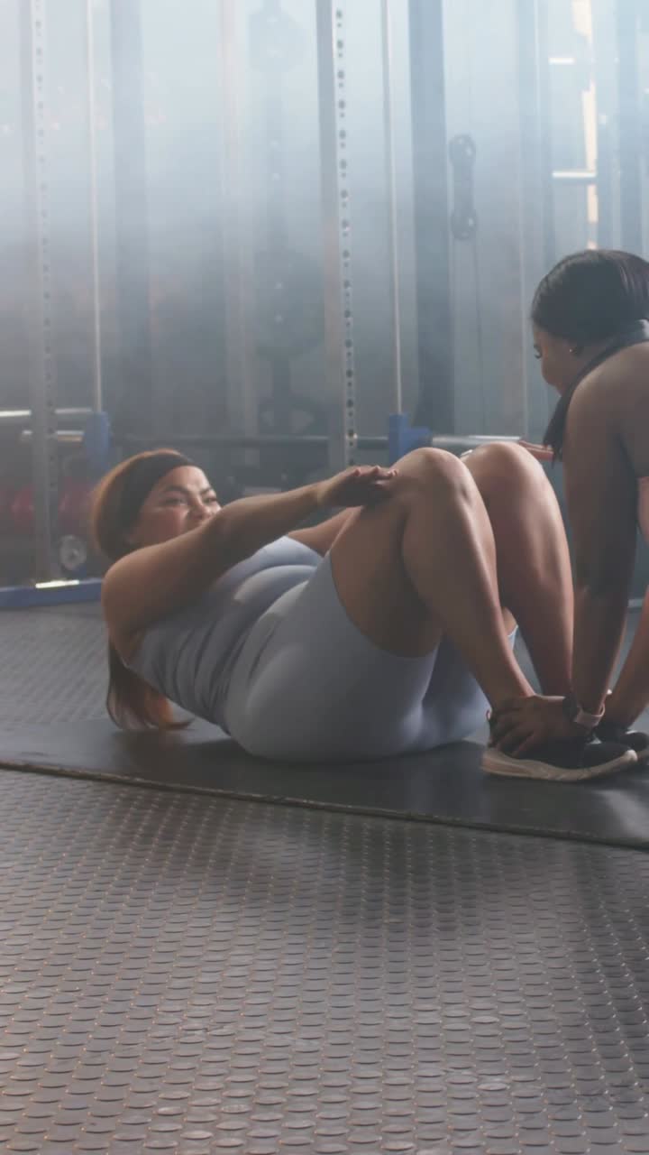 Video verticale: Donna in top grigio che esegue sit-up assistiti sul tappetino per tonificare il core, con un partner che le blocca i piedi.
