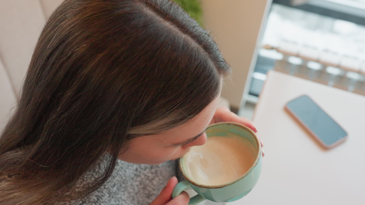 Freelancer in coffee shop drinking coffee with smile lifts head showing happy gestures relaxed atmosphere visible smartphone on table with flower decoration nearby peaceful workspace moment