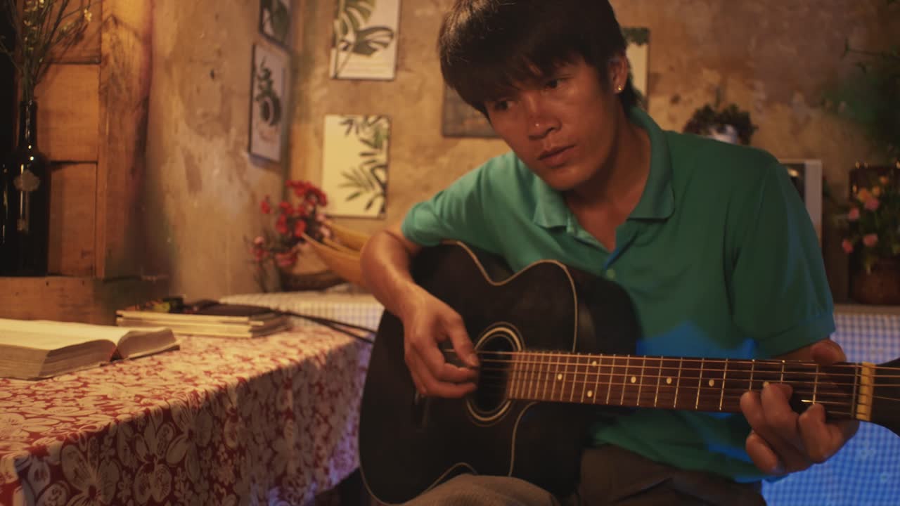 Giovane uomo asiatico che suona la chitarra, seduto su una sedia in una stanza rustica.