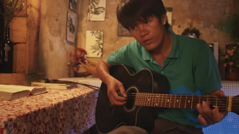 Young-Asian-male-playing-a-guitar-while-sitting-on-a-chair-in-a-rustic-room