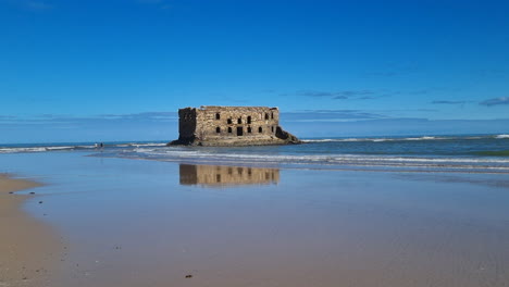 stone ruins of casa del mar reflecting on wet sand of tarfaya beach with ocean waves and blue sky