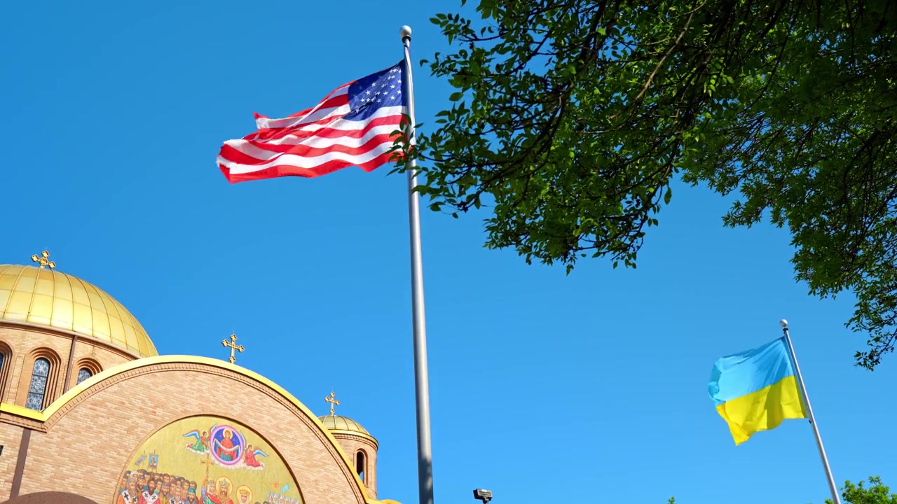 Low angle of flags and gold domed cathedral on a clear day, Chicago. Waving American and Ukrainian banners framed by green tree branches.