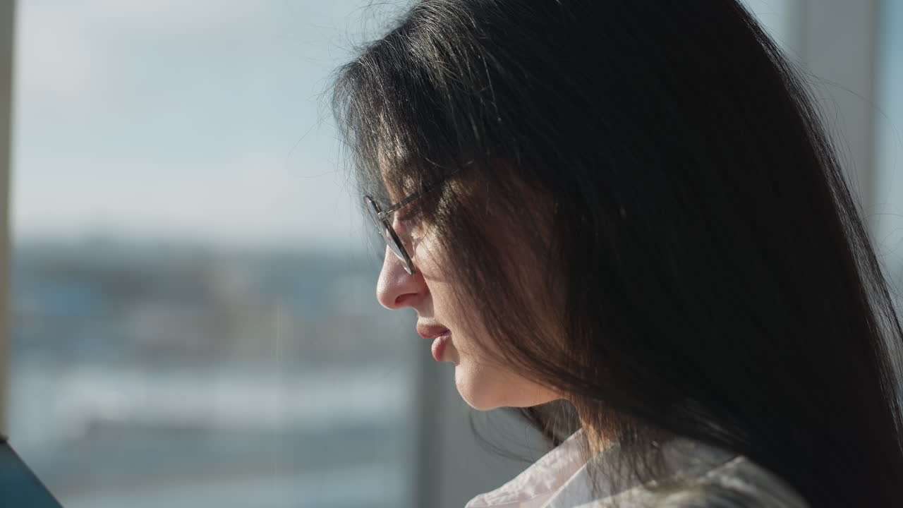 Close up side view of young woman in glasses concentrating on tablet screen indoors, with background featuring soft blur of glass windows and exterior cityscape illuminated by daylight