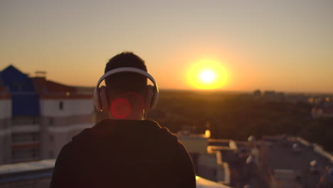 un hombre camina por el techo al atardecer con auriculares mirando a la ciudad desde la altura de un rascacielos al atarrear el sol. relájate mientras escuchas música. disfruta de una hermosa vista de la ciudad al aterrear el sol desde el techo con auriculas