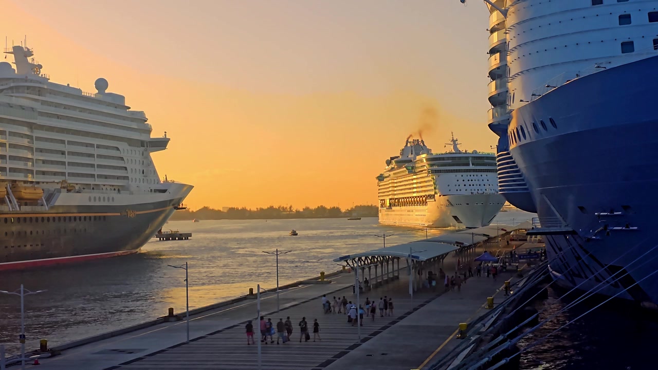 En naturskøn solnedgangsudsigt over et stort Royal Caribbean krydstogtskib, der forlader havnen, indrammet af to andre kolossale, dokkede skibe, mens folk går langs havneterminalens mole.