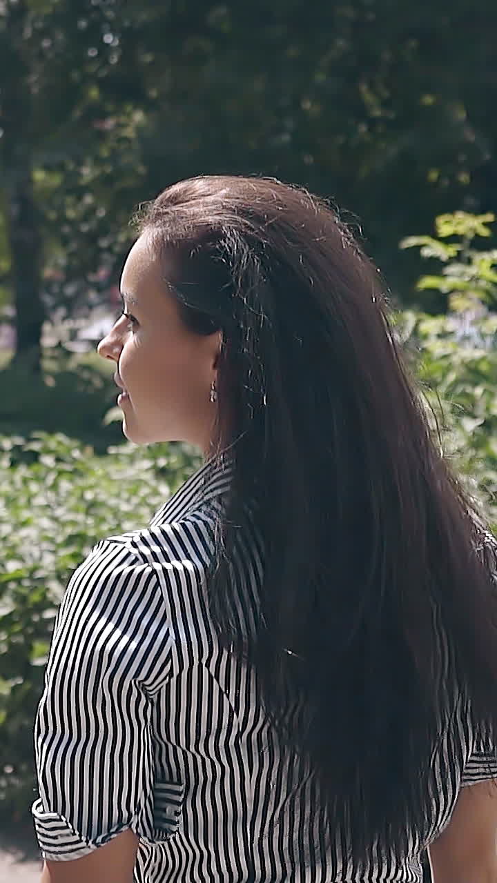 slow motion backside view pretty long haired brunette girl in summer dress with black and white stripes walks in park