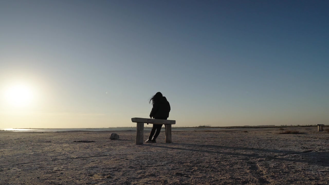 Silhouette posteriore di una donna seduta su una panchina di legno che fissa il cielo deserto all'alba nel Lago Epecuén, Argentina.