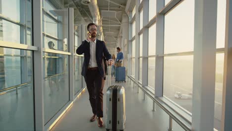 Handsome-Man-In-Business-Style-Stepping-Tothe-Boarding-Through-The-Airport-Corridor,-Carrying-Suitcase-With-Wheels-And-Talking-On-The-Phone,-Businesswoman-Following-Him