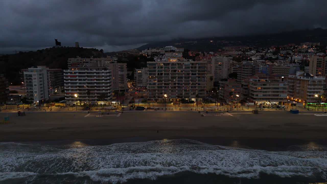 Aerial view capturing Torreblanca beach at dusk, featuring illuminated buildings, crashing waves, and a dark, cloudy sky, all set against the serene Mediterranean coastline in Spain