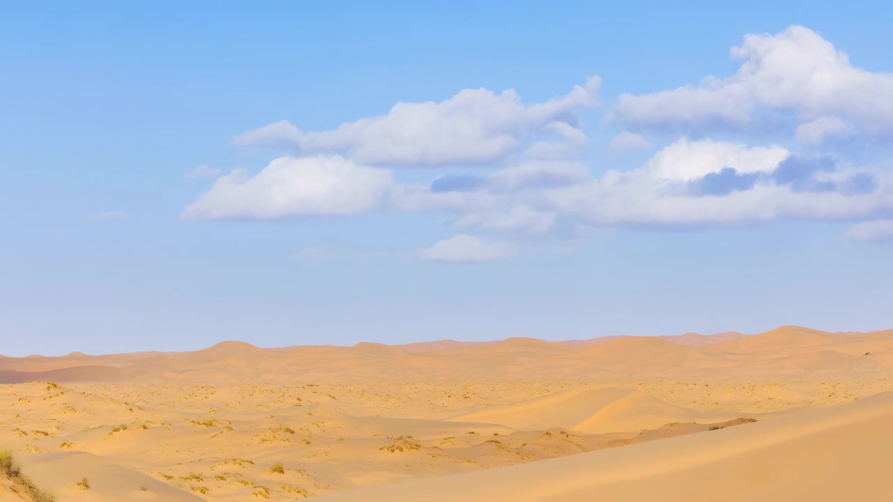 手前から中景にかけて緩やかに起伏する砂丘が広がる、広大な乾燥した風景。散らばる白い雲が浮かぶ明るい青空に縁取られている。