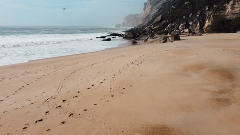 Volando-Sobre-La-Arena-De-La-Playa-Con-Huellas-En-La-Costa-Rocosa-Del-Océano-De-Nazaré-En-Leiria,-Portugal