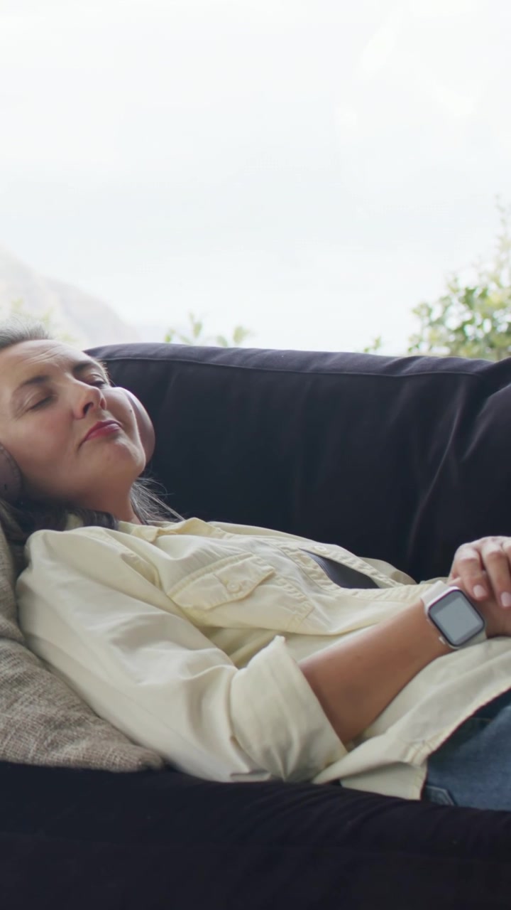 Video verticale: Donna che si sistema sul cuscino, ascolta sul divano vicino alla finestra, con maglietta, cuffie e orologio.