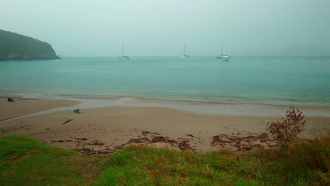 Slow-motion-wide-shot-of-foggy-beach-with-boats-docking-in-the-bay,-at-Urupukapuka-Island,-New-Zealand