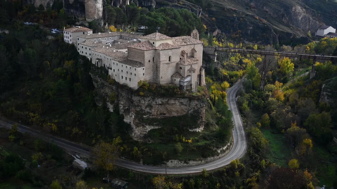 A high-angle aerial view of the Parador de Cuenca in Spain. The historic convent is seen on its cliffside perch above the gorge during a vibrant autumn day.