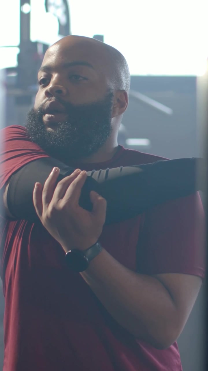 Vertical video: Stepping out behind rack man in maroon tee pulling elbow across chest in gym warmup