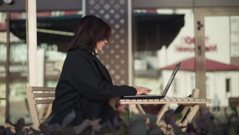 side view of a professional woman typing on laptop outdoors in urban setting, dressed in a chic black coat, with glass building reflecting nearby structures, decorative flowers close to her