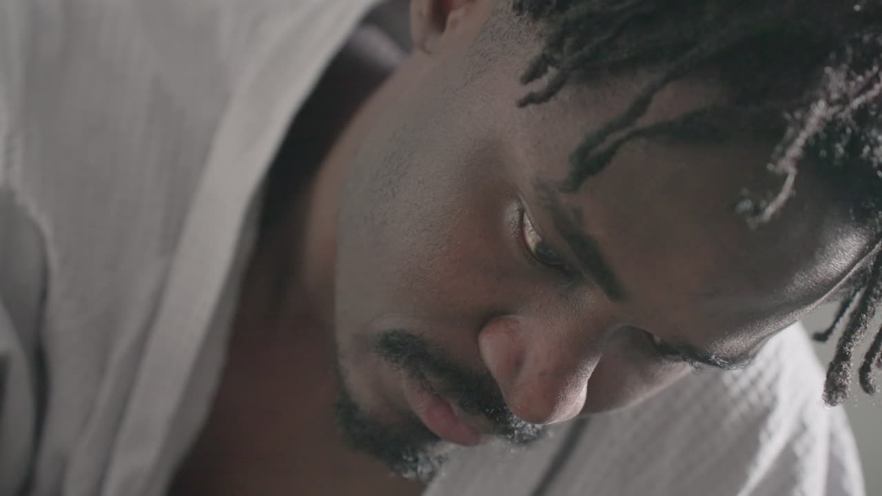 Man in white robe deeply focused while reading large book, expression serious and concentrated, soft natural light from window enhancing peaceful and introspective atmosphere, lying on bed
