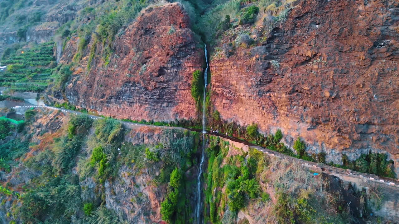 Flying closer to a long thin waterfall falling from a high rock. A group of tourists stands at the highway near the water. The Madeira Islands, Portugal.