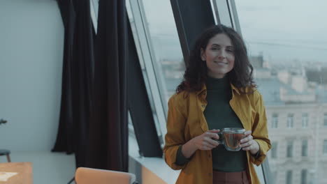 Happy-Woman-Holding-Tea-Cup-and-Smiling-at-Camera-in-Office