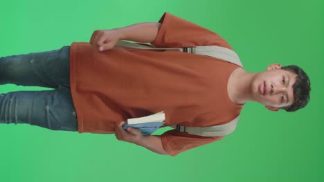 the front view of asian boy student wearing a backpack, brown t-shirt, and blue jeans holding a book and walking to school on green screen chroma key.