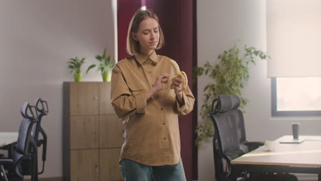 Pregnant-Happy-Woman-Standing-Eating-A-Banana-And-Looking-At-Camera-In-The-Office