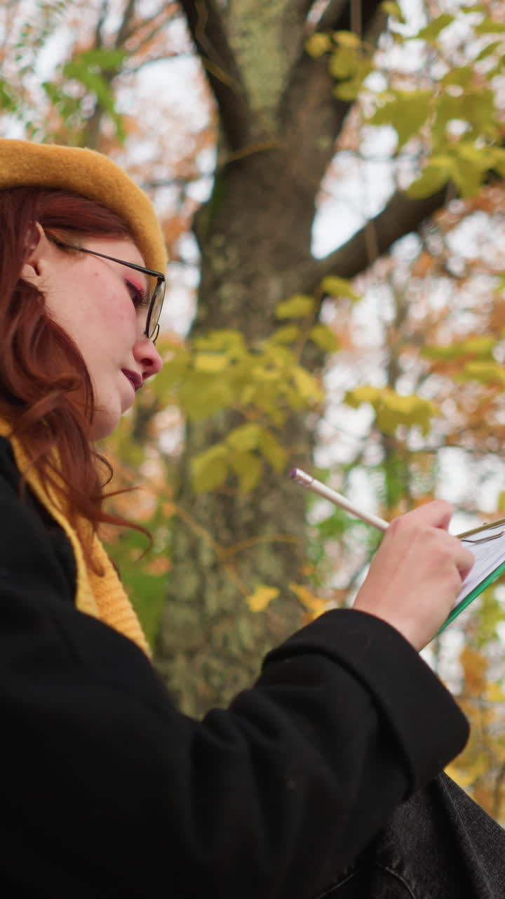 선글라스, 노란 베레트, 노란 마플러를 입은 창의적인 여성의 측면 뷰, 그녀의 그림에 깊이 집중, 활기찬 가을 잎으로 둘러싸인 벤치에 앉아