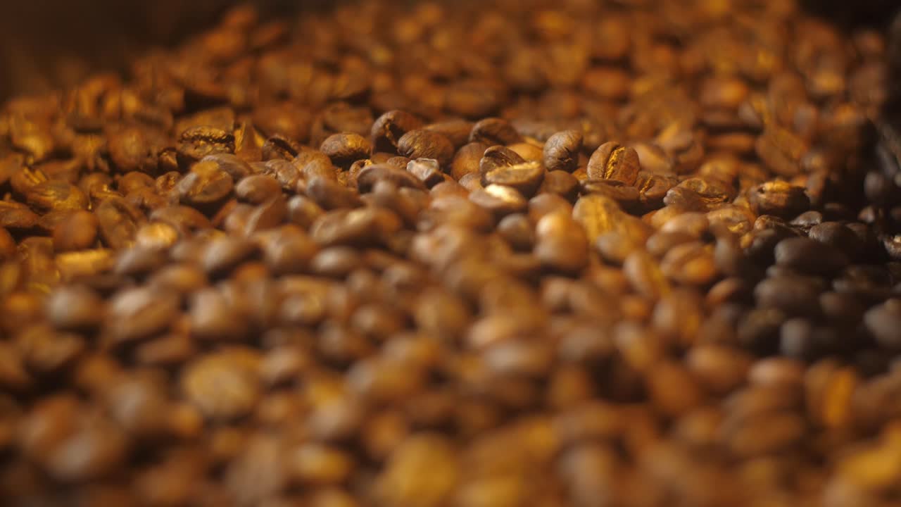 Close-up de uma pilha de grãos de café torrados em um tom médio-escuro. Os grãos têm uma textura fosca e são iluminados por uma luz quente, sugerindo frescor e aroma.