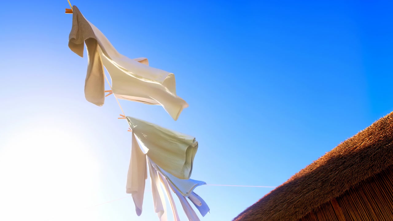 Tessuto bianco pende in primo piano su uno stendibiancheria, risplendendo sotto la forte luce del sole contro un cielo azzurro limpido, con un tetto di paglia rustico visibile a destra