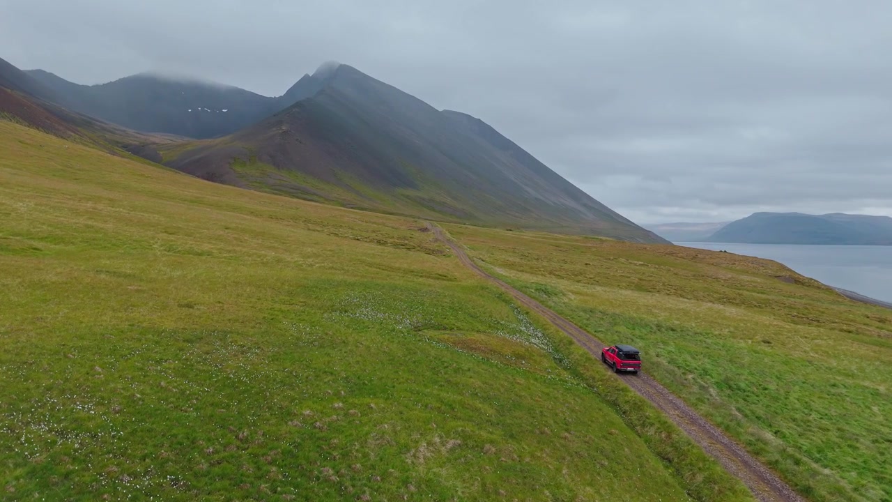 Ripresa aerea cinematografica di un veicolo 4x4 rosso che viaggia lungo una remota strada sterrata nei Fiordi Occidentali dell'Islanda, con montagne spettacolari e paesaggi costieri.