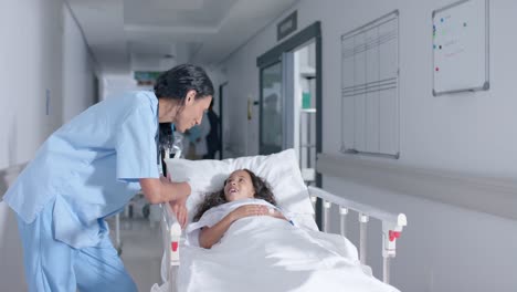 diverse female doctor and child patient talking in corridor at hospital, in slow motion