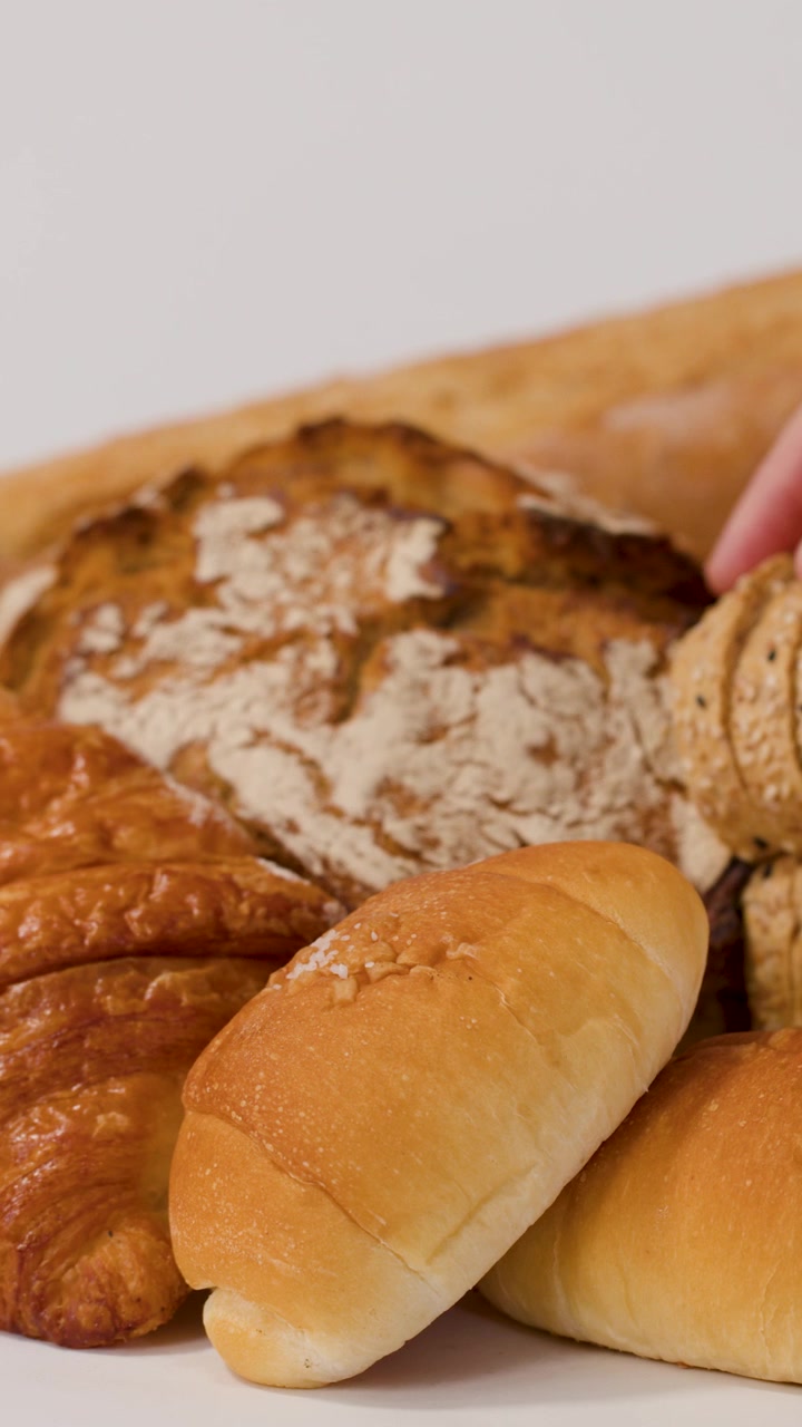 Close-up of hands placing sliced whole grain bread among croissants and baguettes on white.