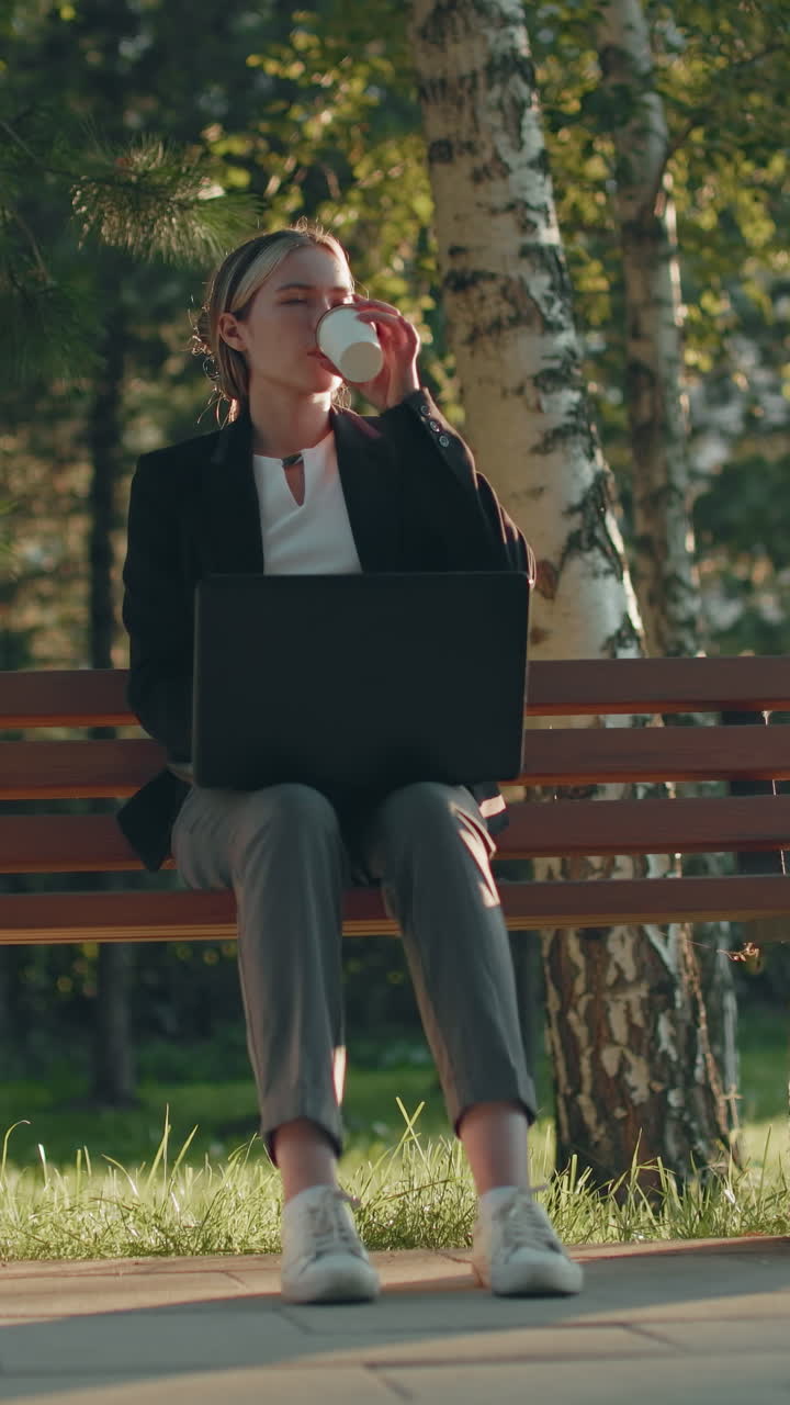 Female freelancer seated on wooden bench outdoors working with laptop on lap, taking break to drink water, surrounded by lush trees, grass, and sunlight in peaceful green park setting