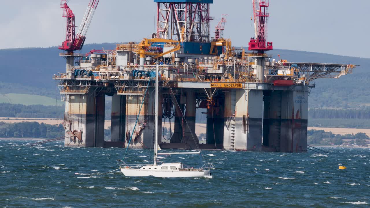 Hvid sejlbåd passerer stor offshore olieplatform på oprørt hav, dagslys, bredt billede.