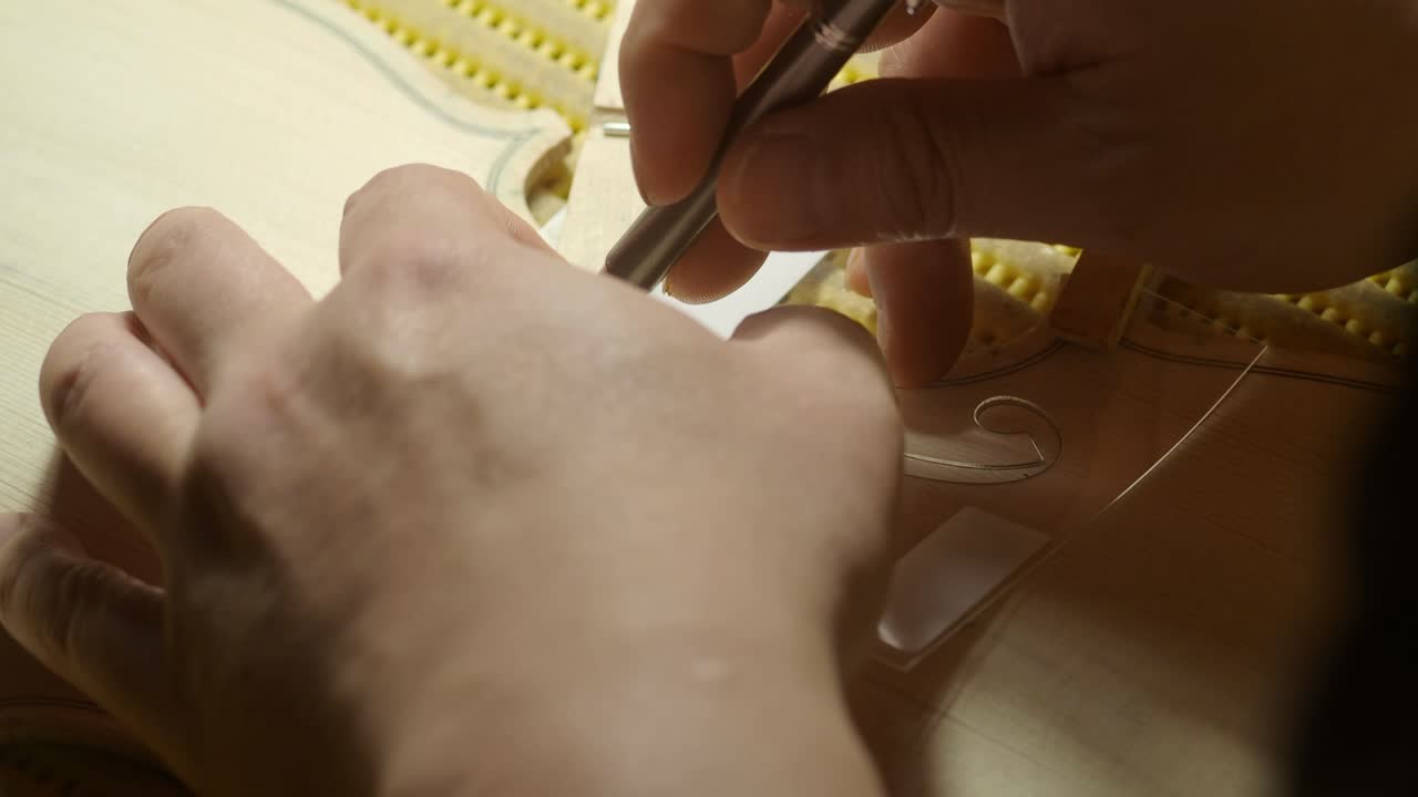 Skilled luthier carefully marking f hole outline on violin top plate soundboard, using wooden template and pencil in time honored woodworking workshop, real time