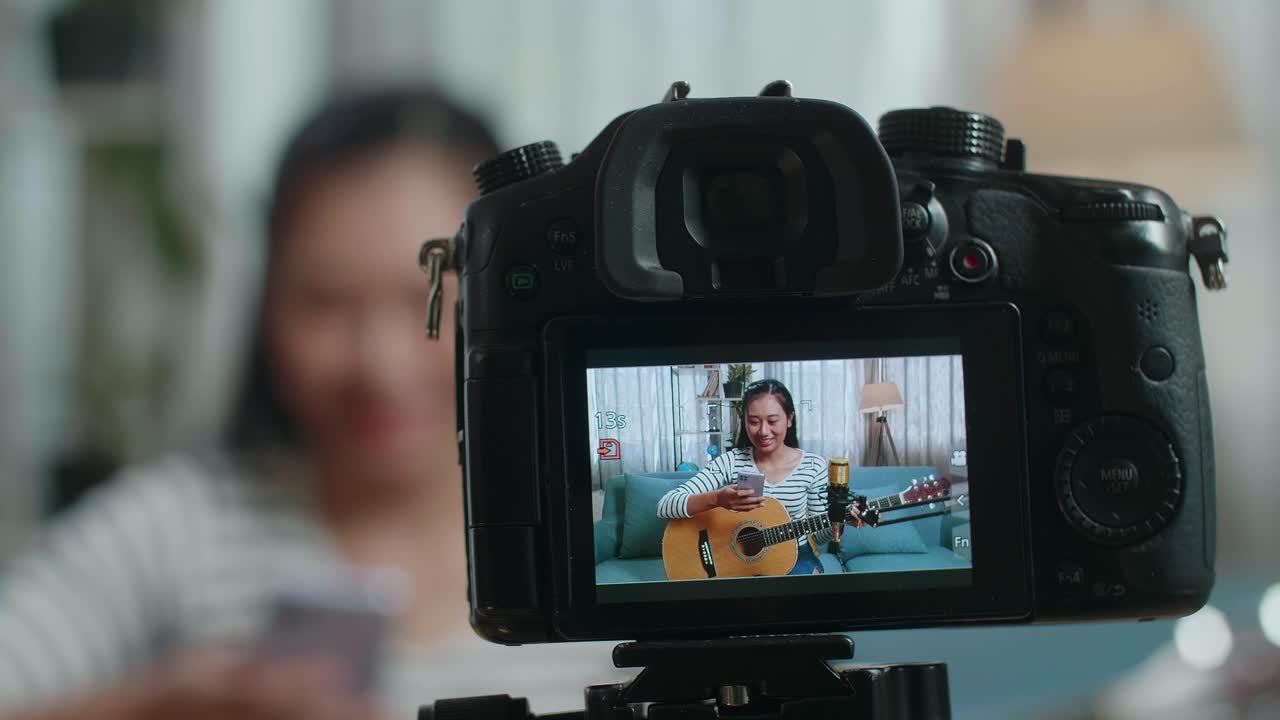 Närbild av en kameramonitor som spelar in asiatisk kvinna som läser kommentarer på smartphone och pratar medan hon livestreamar och spelar gitarr hemma