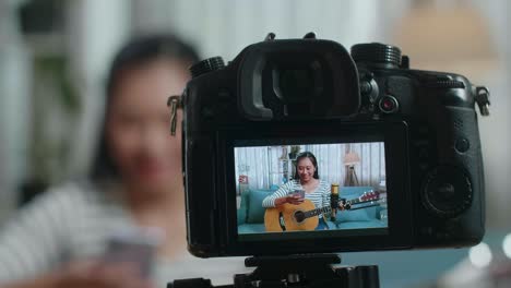 close up of a camera monitor recording asian woman reading comments on smartphone and speaking while live stream playing guitar at home